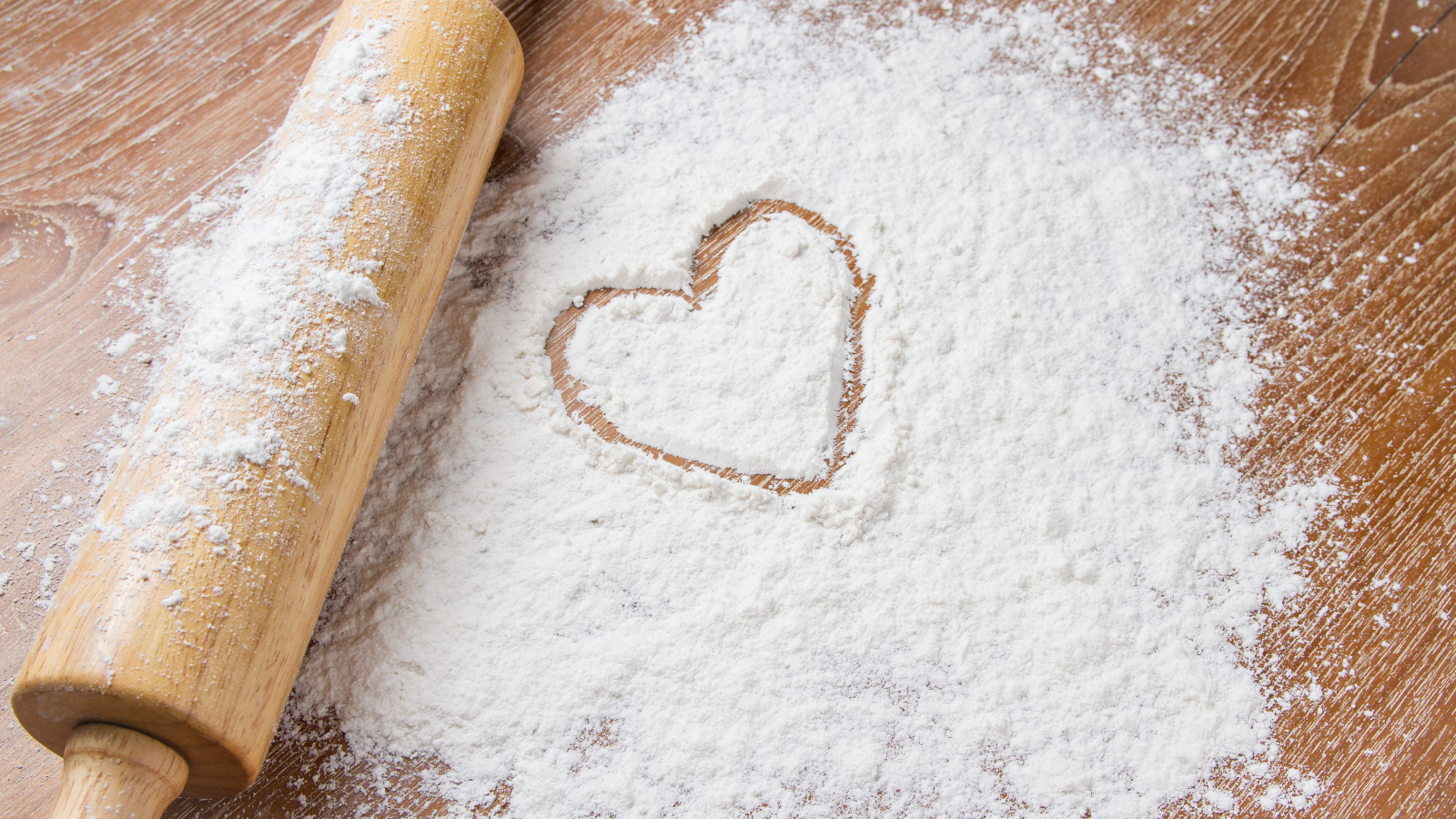 Image of a rolling pin rolling out flour with a love heart shape drawn in the flour to illustrate making mince pies